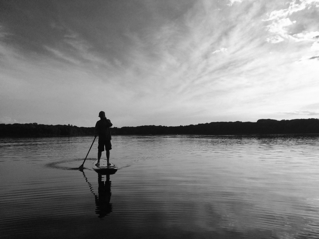 man on a handmade wooden stand up paddle board on a lake in black and grey photography
